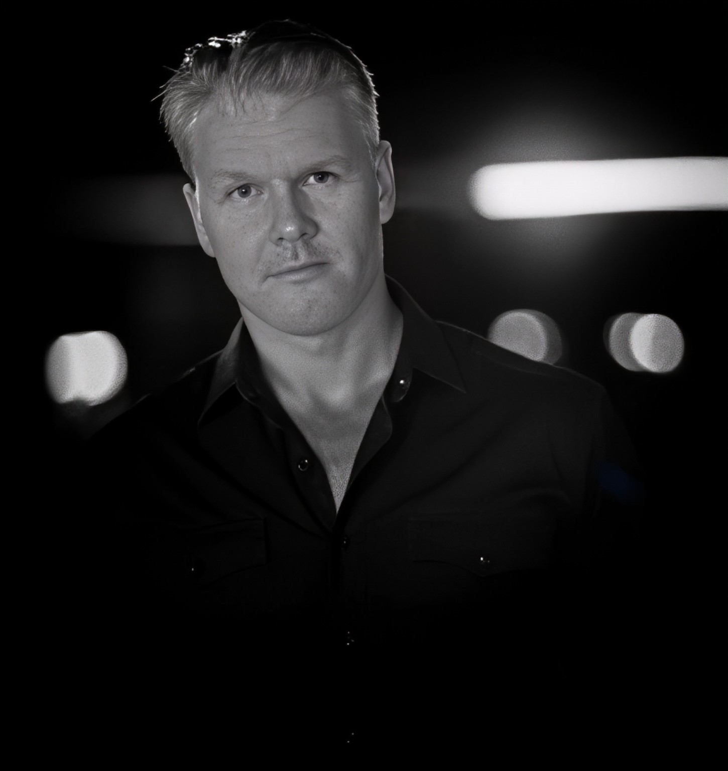 Black and white portrait of Andrew Pacholyk in a dark shirt, with stage lights in the background.